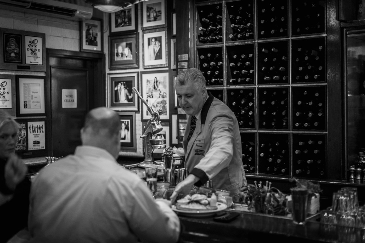 A bartender with gray hair serves food to a bald customer at a bar. The bar has a wall of wine bottles and framed photos. The scene is in black and white, creating a classic, nostalgic atmosphere.