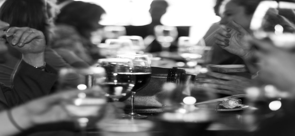 A black and white photo of a social gathering. Several people are seated at a table with glasses of wine and other drinks. The foreground shows blurred hands holding glasses, with a bag visible on the table. The background is out of focus.