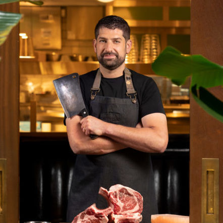 A man wearing a black shirt and apron stands in a kitchen, holding a large meat cleaver. In front of him is a table with raw cuts of meat. The background has kitchen equipment and potted plants on either side.