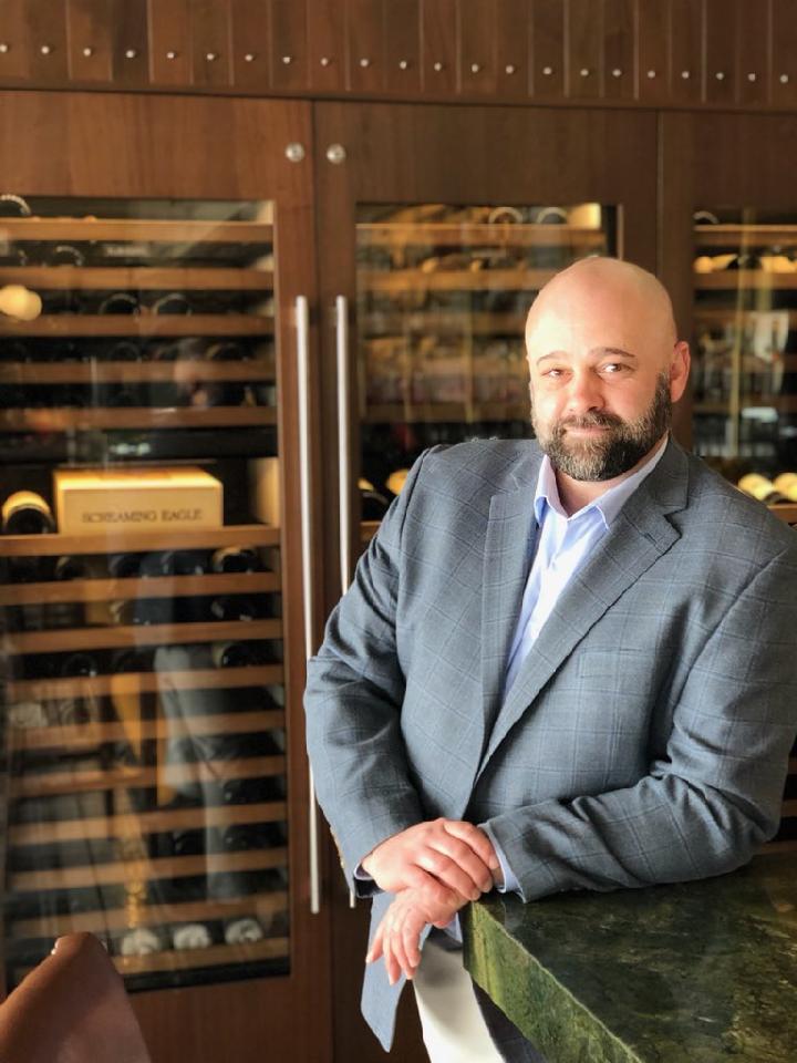 A man with a beard and shaved head, wearing a gray suit jacket and white shirt, leans against a green marble counter. Behind him, there is a wooden wine cabinet with several bottles visible.