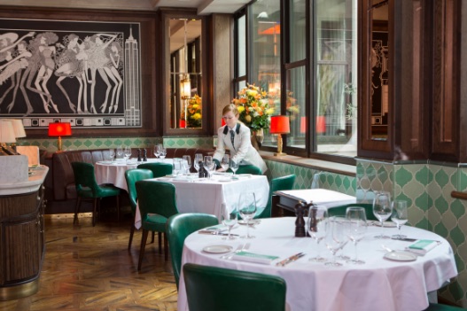 Stylish restaurant interior with a server setting a table. The room features green chairs, tables with white cloths, and elegant decor, including a black and white mural on the wall. Bright natural light comes through large windows.