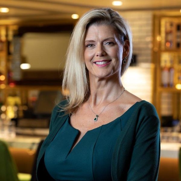 Smiling woman with long blonde hair wearing a teal dress and necklace, standing indoors in a warmly lit setting with blurred background.