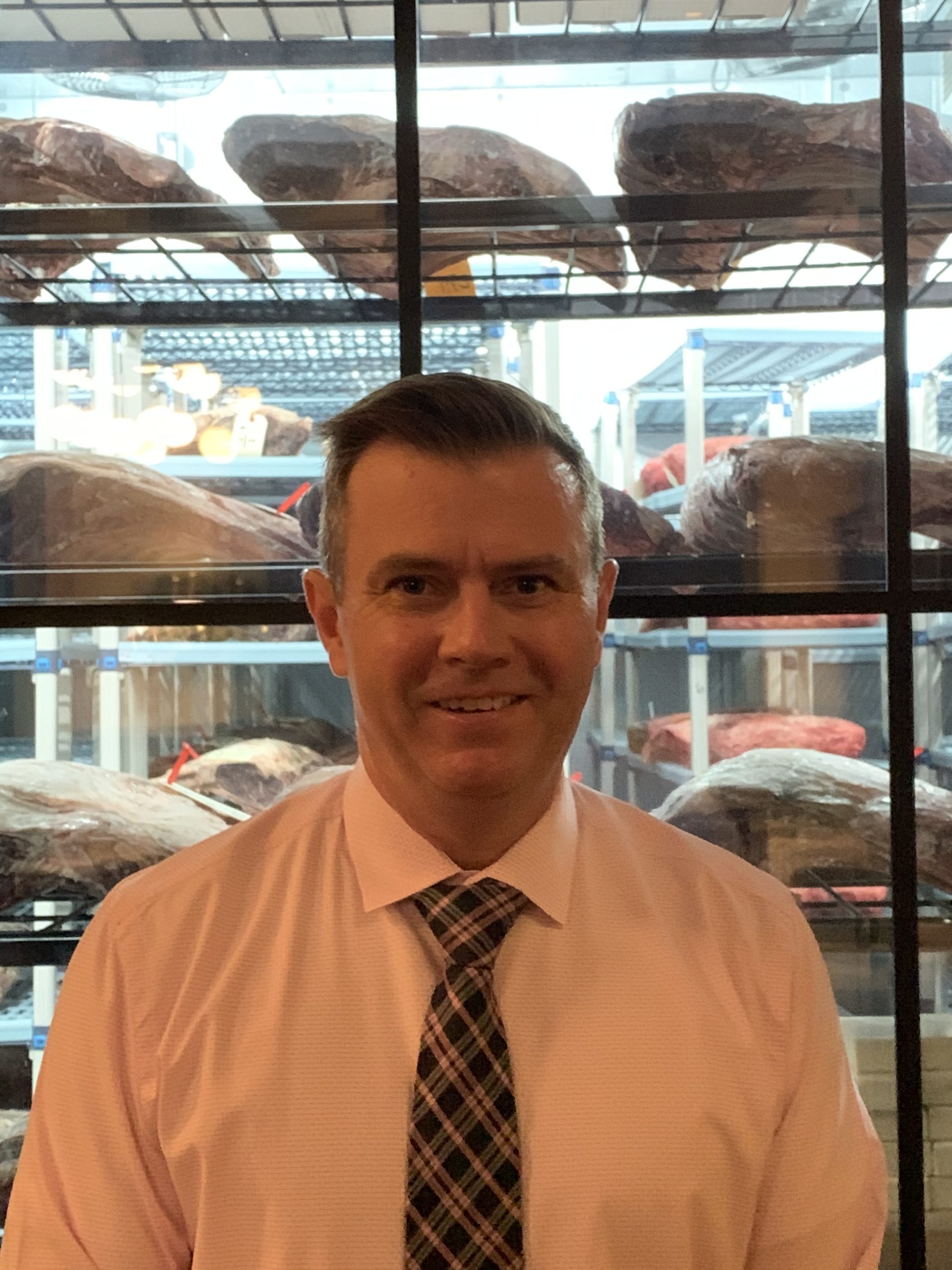 A man in a pink shirt and plaid tie stands smiling in front of shelves displaying large cuts of meat. The shelves are behind glass, and the setting appears to be a butcher shop or meat storage area.