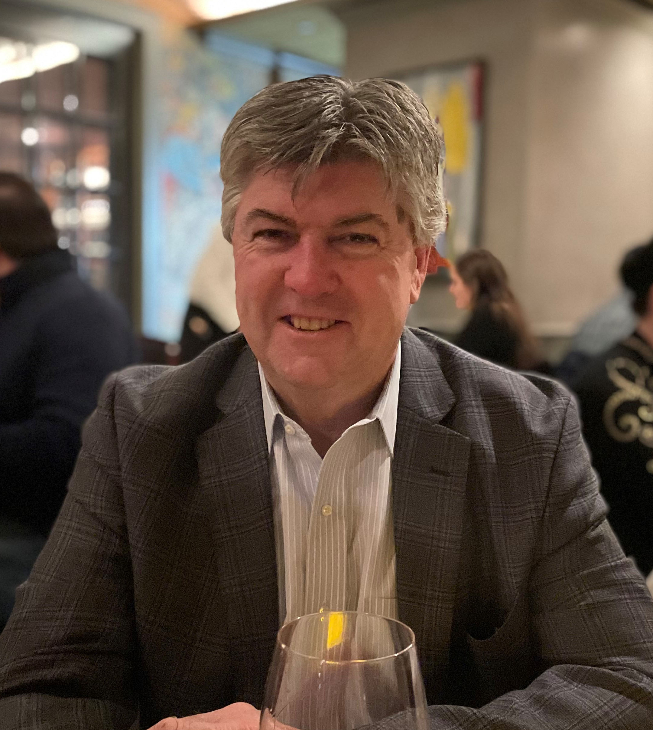 A man with gray hair is sitting at a table in a restaurant, smiling at the camera. He is wearing a checkered blazer and a striped shirt. A wine glass is on the table in front of him. The background shows other diners and blurred decor.