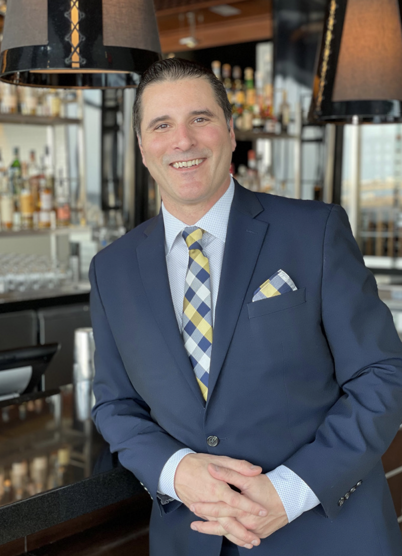 Smiling man in a dark suit with a checkered tie and matching pocket square stands in a bar area. Bottles of alcohol are visible on shelves in the background. He has short dark hair and is leaning casually on the bar counter.