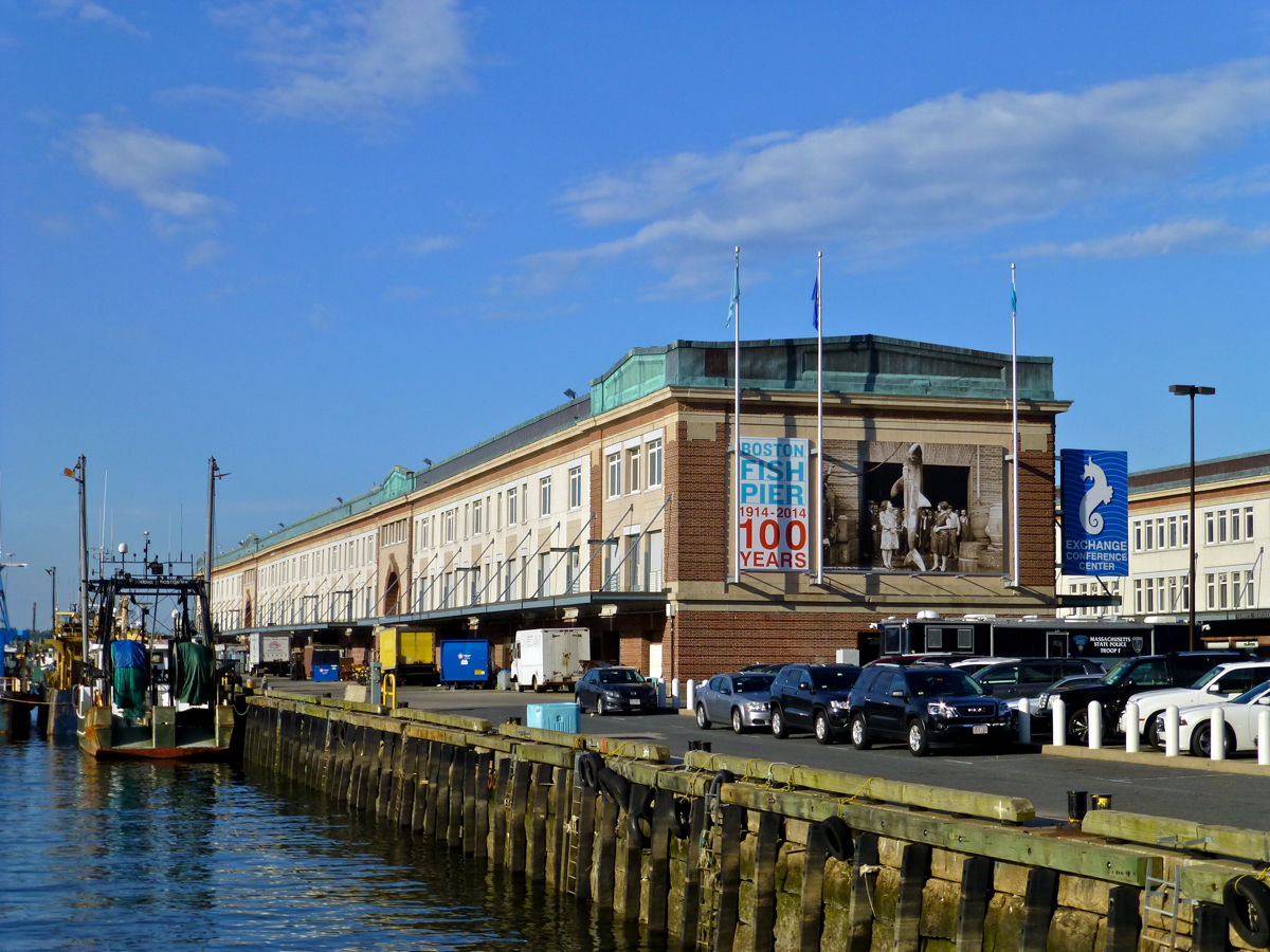 View of the Boston Fish Pier, a large waterfront building with a historic sign celebrating 100 years. Several cars are parked in front, and a few fishing boats are docked at the pier. The sky is blue with some clouds.