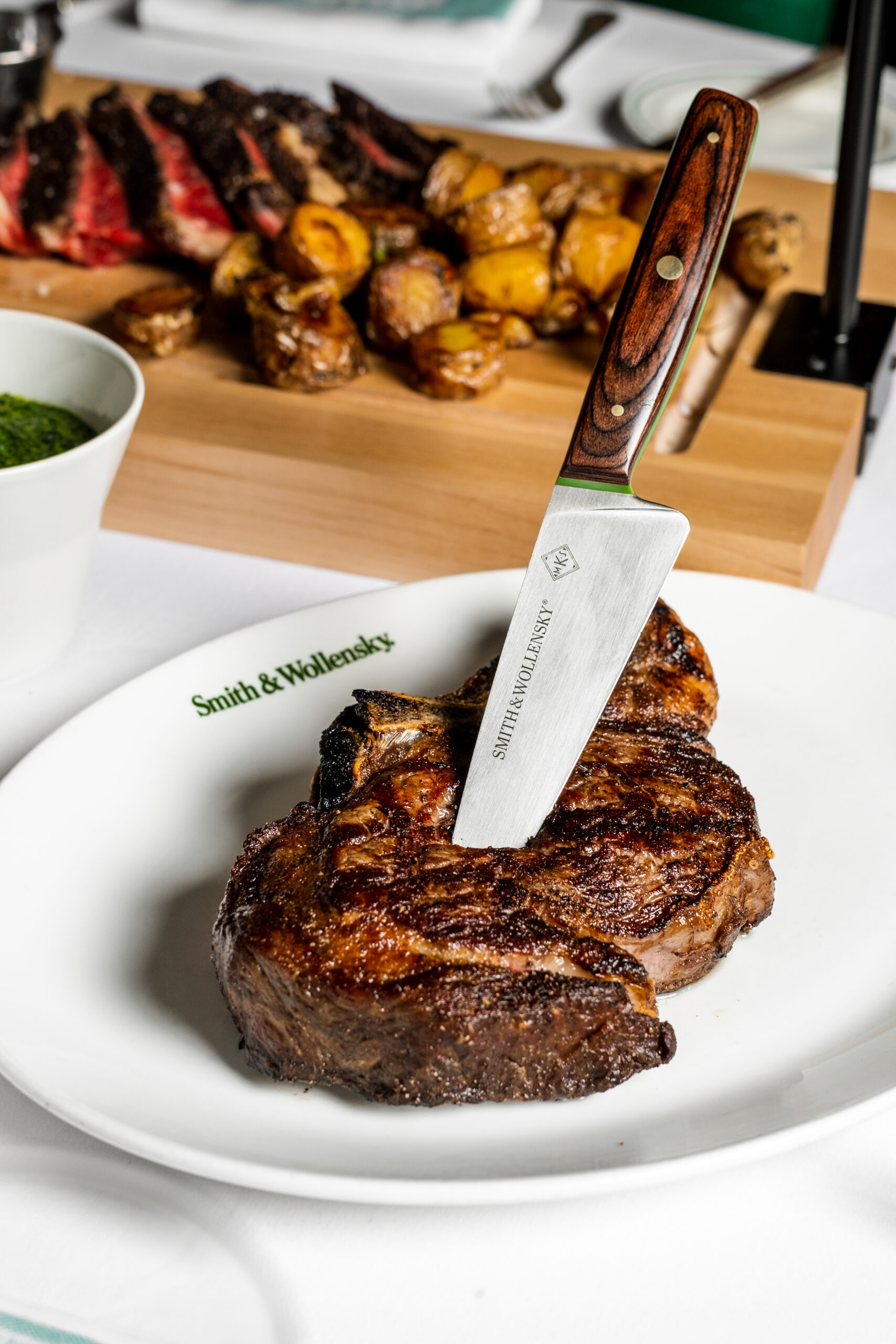 A grilled steak with a knife stuck in it is served on a white Smith & Wollensky plate, with roasted potatoes and sliced meat on a wooden board and a bowl of green sauce in the background.