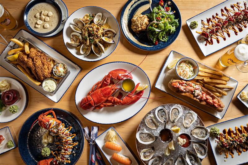 A variety of seafood dishes on a wooden table, including oysters, lobster, clams, sushi, fish and chips, salad, and drinks, all artfully arranged on plates.